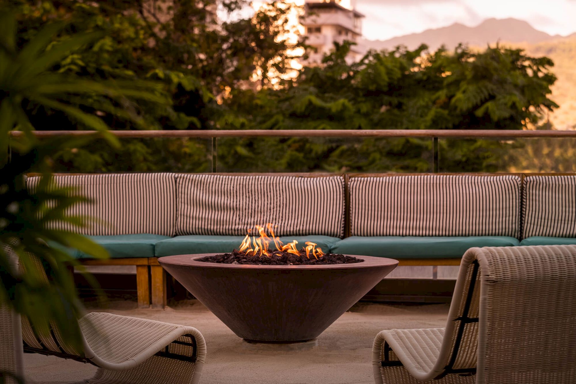 An outdoor seating area with a fire pit in the center, surrounded by wicker chairs and lush greenery in the background.