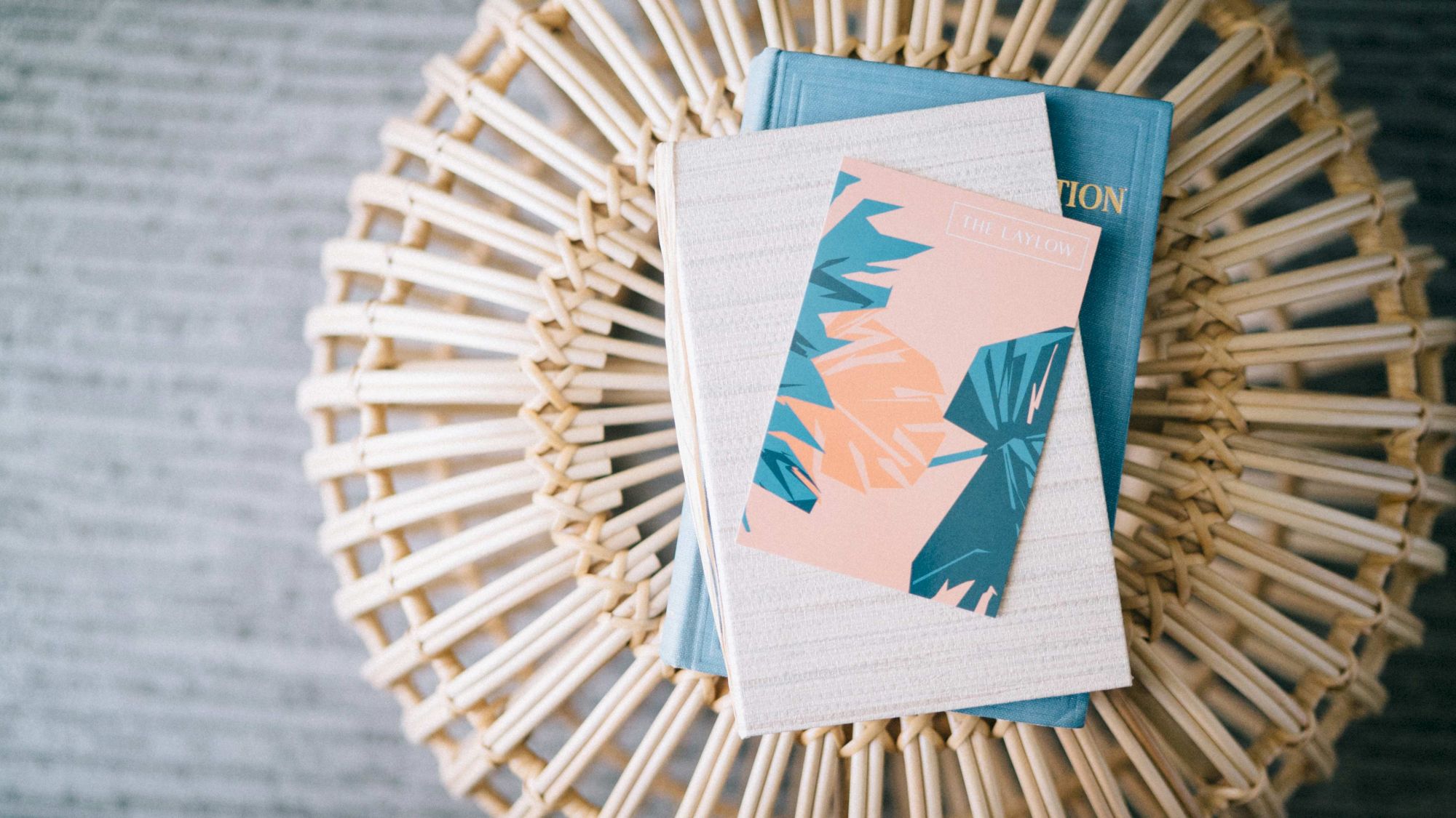 A wicker stool holds a stack of books, with the top book featuring a pink and blue leaf design cover.