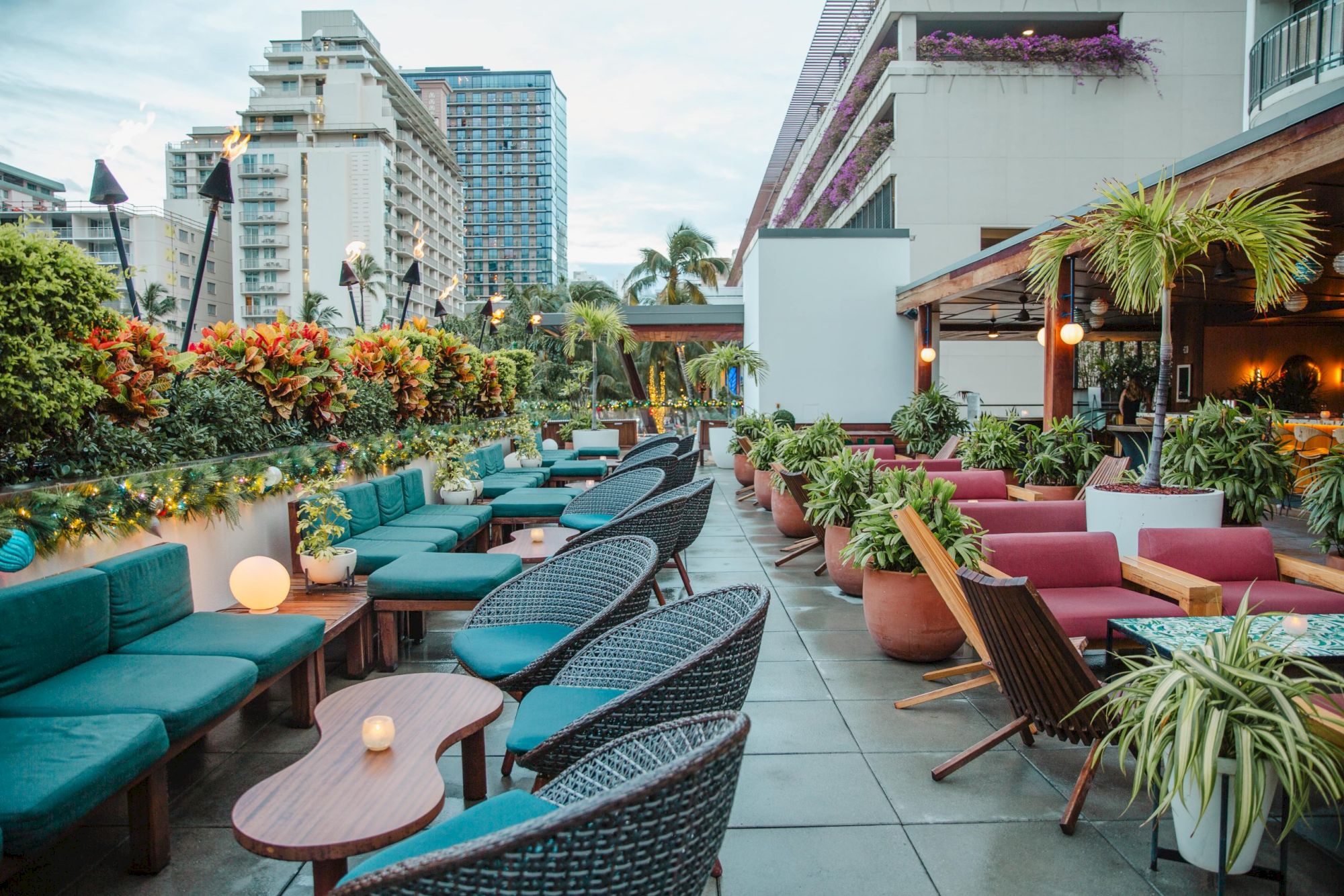 Rooftop terrace with cozy seating, lush plants, and ambient lighting, surrounded by buildings and greenery.