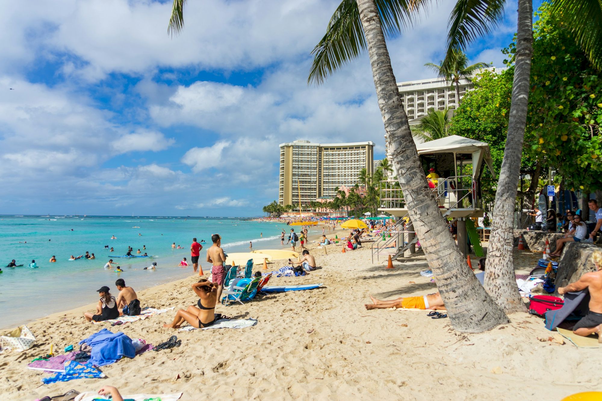 People relaxing on a beach with palm trees, umbrellas, and a hotel in the background, enjoying a sunny day by the ocean.
