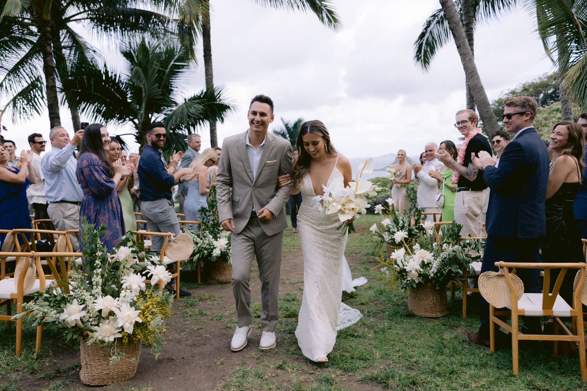 A bride and groom walk down an outdoor aisle surrounded by guests applauding, with tropical plants and palm trees in the background.