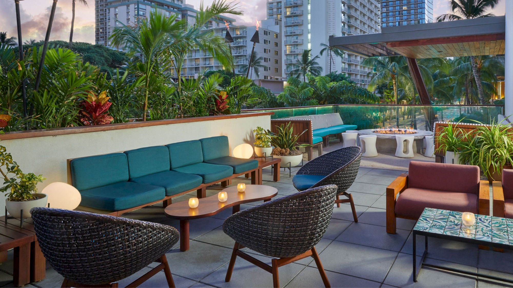 Rooftop patio with modern seating, tables, plants, and candles. Cityscape in the background. Cozy atmosphere for relaxation or gatherings.