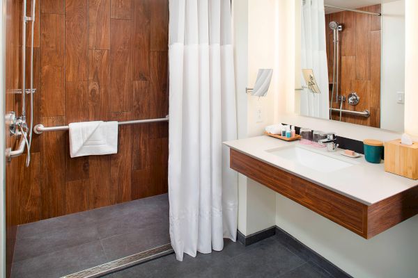 The image shows a modern bathroom with a walk-in shower, white curtain, wooden accents, sink, and mirror.