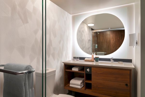A modern bathroom with a circular mirror, wooden vanity, and neatly arranged towels and toiletries, illuminated by soft lighting.