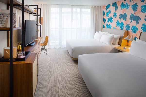 A hotel room featuring two beds, a tropical-themed accent wall, a TV on a stand, a desk with a chair, and large windows with sheer curtains.