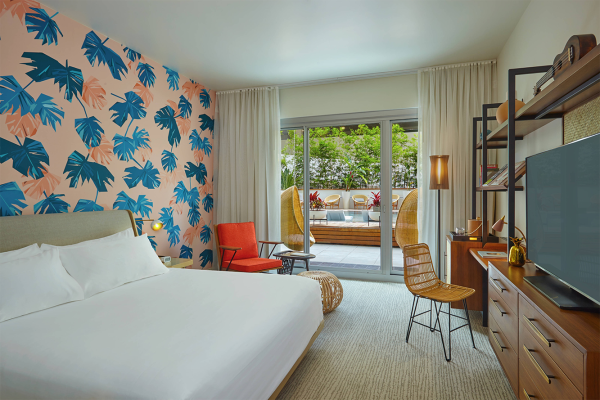 The image shows a modern bedroom with tropical-themed wallpaper, a bed, a TV, and a view of an outdoor patio through sliding doors.