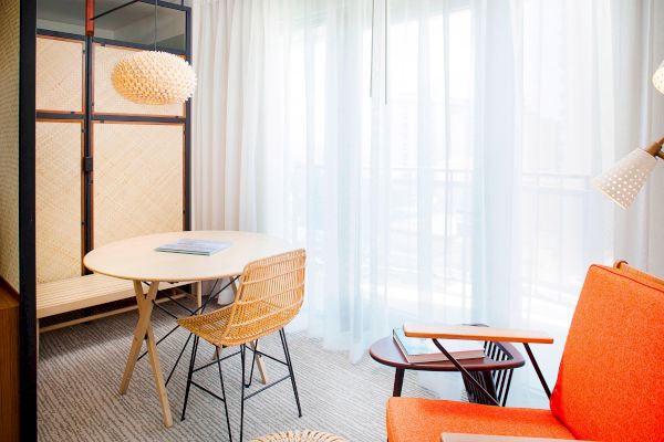 The image shows a modern room with an orange armchair, round table, wicker chair, and a woven pendant light near sheer curtains.