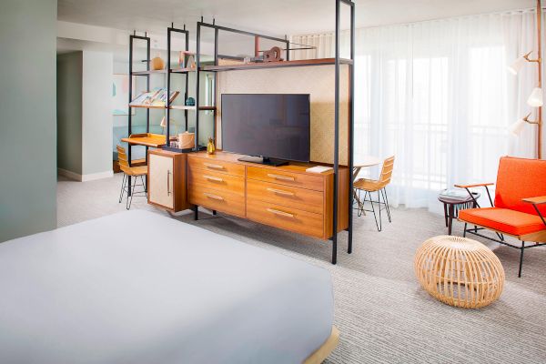 The image shows a modern hotel room with a bed, TV, seating area, shelves, and a window with sheer curtains.