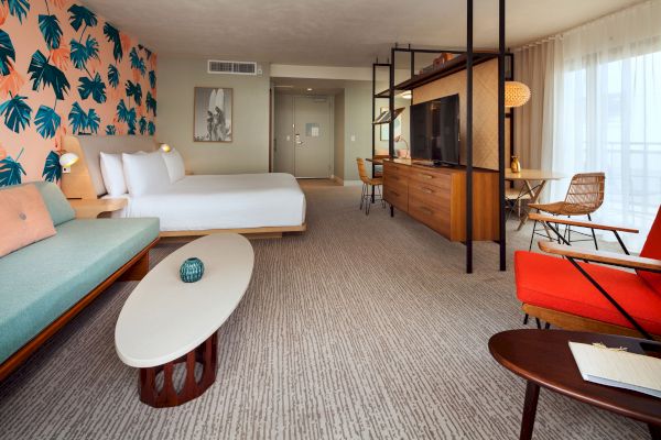 The image shows a modern hotel room with a double bed, colorful furnishings, a seating area, and a TV on a stand with a partition.