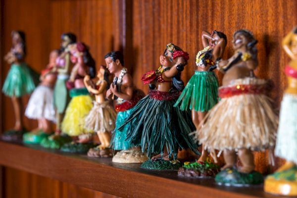 A row of colorful hula dancer figurines with grass skirts is displayed on a wooden shelf, each in varied poses.