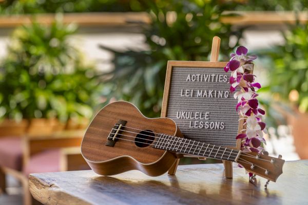 A ukulele rests on a table beside a sign with activities: "Lei Making" and "Ukulele Lessons," adorned with orchids.