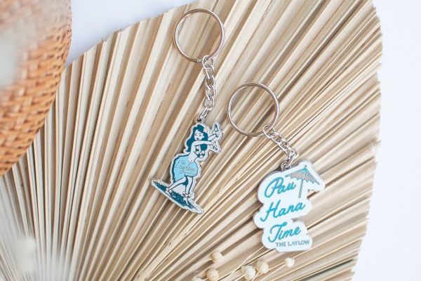 Two keychains are on a woven, fan-shaped surface. One has a blue character, the other has text saying "It's Hanu Time" with a small surfboard.