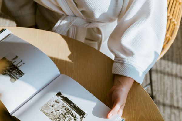 A person in a white robe is sitting at a table, holding an open book with black-and-white photographs, with sunlight casting shadows.