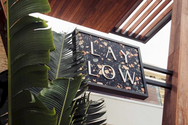 The image shows a sign reading "LAY LOW" with a terrazzo design, next to large green leaves and wooden architectural elements.