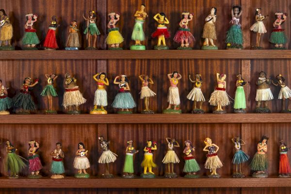 The image shows a collection of miniature hula dancer figurines in various poses, displayed on wooden shelves.
