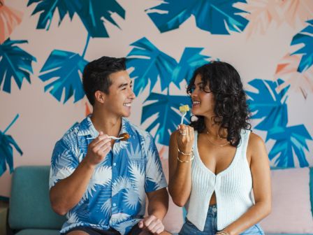 A couple sits on a couch with a tropical leaf pattern background, enjoying food and drinks, and smiling at each other.
