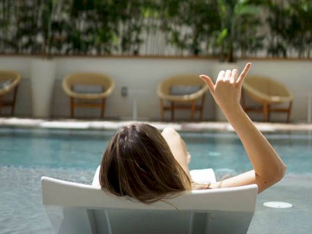 A person relaxes on a poolside lounge chair, gesturing. There are chairs and plants in the background, suggesting a tranquil setting.