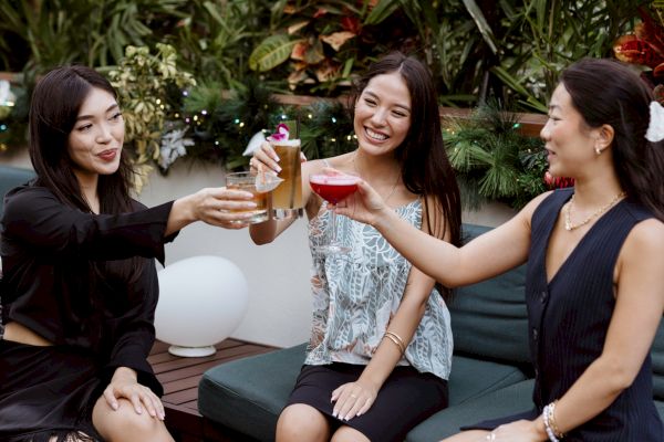 Three people are sitting outside, toasting with drinks and smiling, surrounded by lush greenery and a festive atmosphere.