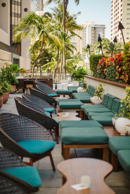 Outdoor seating area with wicker chairs and green cushions, surrounded by plants and cityscape in the background.