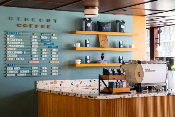 The image shows a coffee shop counter with a price board, bags of coffee, cups, a coffee machine, and wooden shelves on a blue wall.