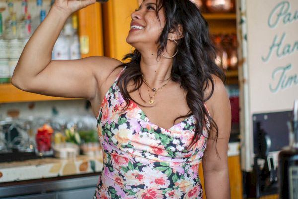A woman in a floral dress is smiling while shaking a cocktail shaker in a bar setting.