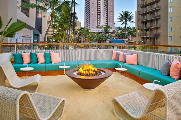 An outdoor lounge with a fire pit, surrounded by teal and pink cushioned seating, palm trees, and city buildings in the background.