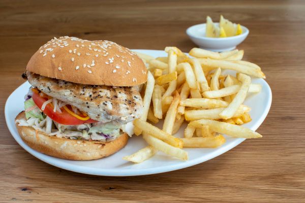 A chicken sandwich with lettuce and tomato, served with a side of french fries, and a small bowl of lemon slices on a wooden table.