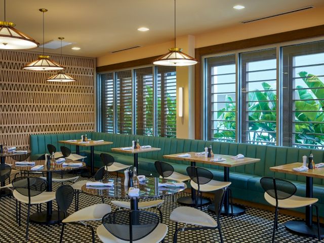 A stylish restaurant interior with patterned floors, green seating, wooden tables, and modern lighting near large windows with leafy views.