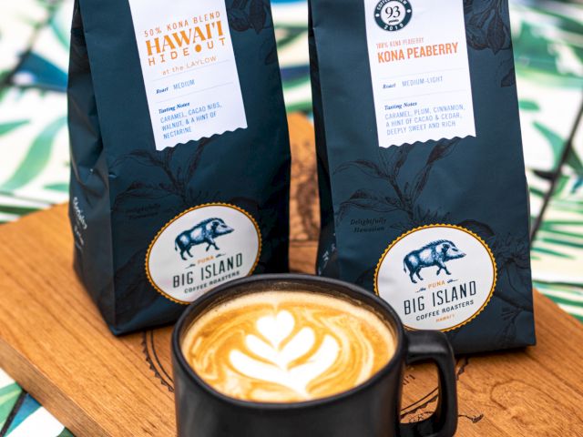 A mug of latte with latte art sits on a wooden board beside two bags of coffee. The background has a green, leafy design pattern.