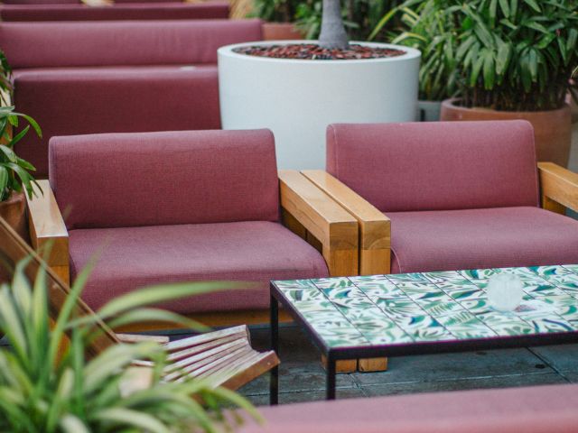 Outdoor seating area with wooden couches, red cushions, and a glass-top table surrounded by lush green plants and potted plants.