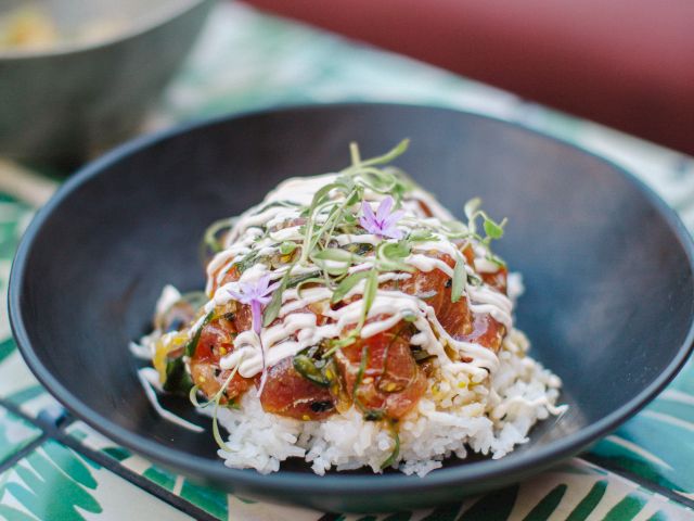 A black bowl with rice topped with diced vegetables, sauce, and herbs on a patterned table.