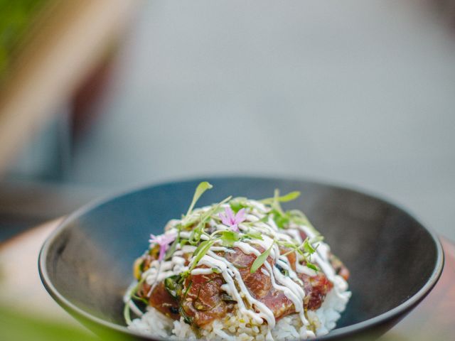 A dish in a black bowl featuring rice topped with meat, sauce, and greens, garnished with sprouts in a blurred outdoor setting.