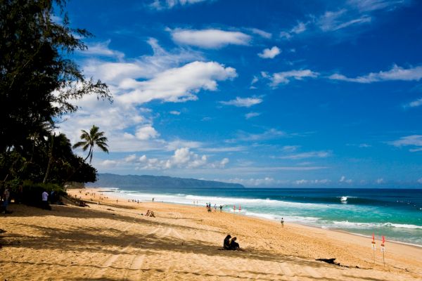 A sunny beach scene with golden sand, a few scattered people, palm trees, and a vibrant blue ocean under a sky dotted with clouds.