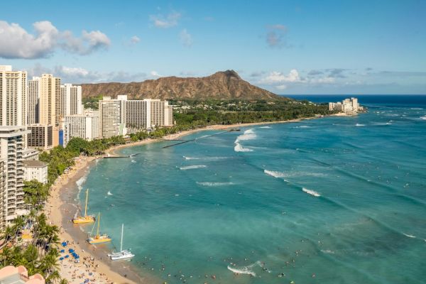 A beach with turquoise water, high-rise buildings, a lush mountain in the background, and boats along the shoreline.