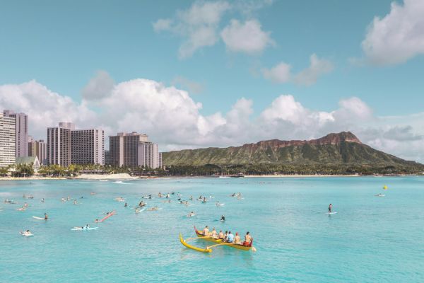 A vibrant beach scene with clear blue water, people kayaking and paddleboarding, skyscrapers, and a mountain in the background.