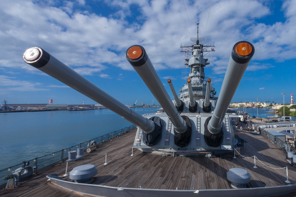 A battleship is shown with large artillery guns on its deck, docked in a harbor under a partly cloudy sky.