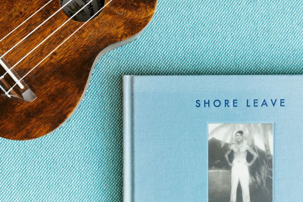 A ukulele lies next to the book "Shore Leave" by Ryan Mungia, featuring a vintage photograph on a blue textured background.