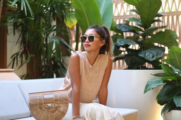 A person is sitting on an outdoor sofa surrounded by lush green plants, wearing sunglasses and casual attire, with a woven bag beside them.