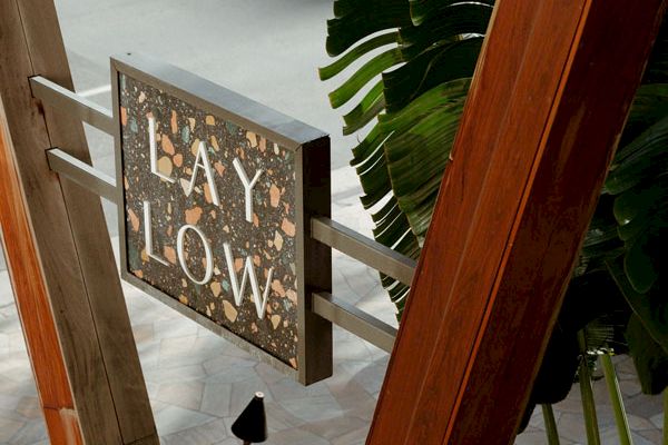 The image shows a sign with the words "LAY LOW" mounted on a structure, surrounded by wooden beams and tropical plants.