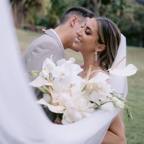 A couple embraces on their wedding day, outdoors with lush greenery. The bride holds a bouquet of white flowers, veil flowing.
