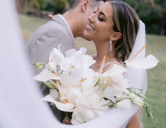 A couple embraces on their wedding day, outdoors with lush greenery. The bride holds a bouquet of white flowers, veil flowing.