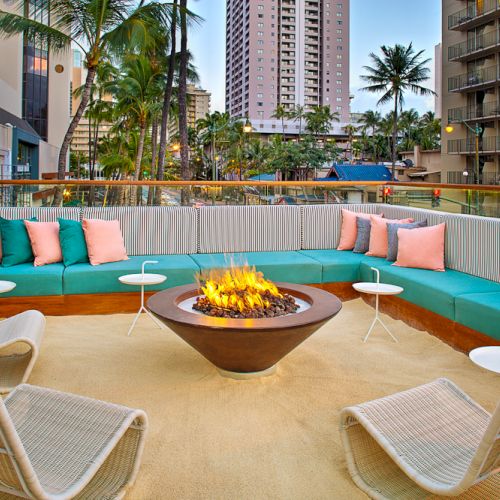 An outdoor lounge with turquoise seating, a fire pit, and palm trees, surrounded by city buildings at dusk.