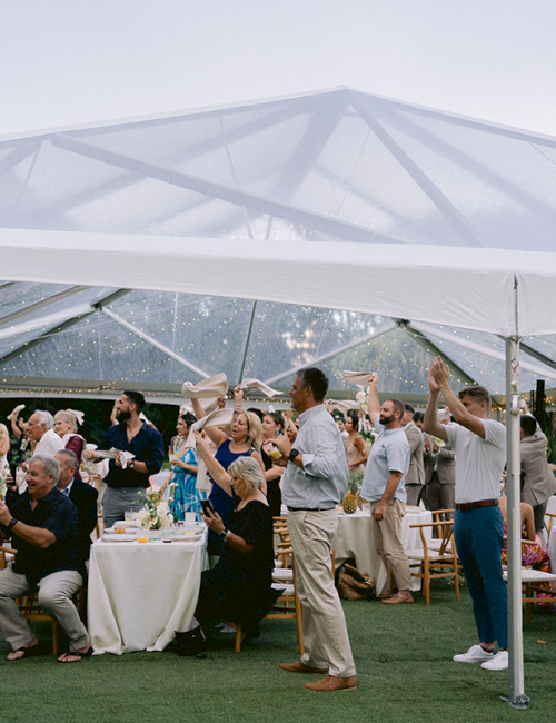 A group of people are celebrating under a clear tent, with some raising glasses in a festive outdoor gathering.