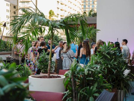 A group of people socialize in a lush, plant-filled outdoor setting, possibly a garden or rooftop event space.