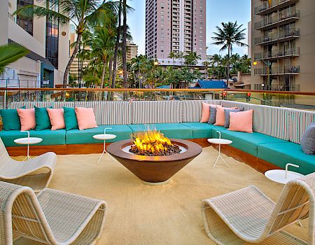 A cozy outdoor seating area with a fire pit, surrounded by vibrant cushions and buildings, offers a relaxing urban retreat.