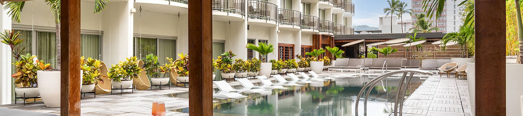 A serene hotel pool area with lounge chairs, cabana, and tropical plants under a blue sky, perfect for relaxing.