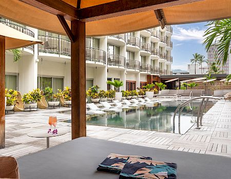 A serene hotel pool area with lounge chairs, cabana, and tropical plants under a blue sky, perfect for relaxing.