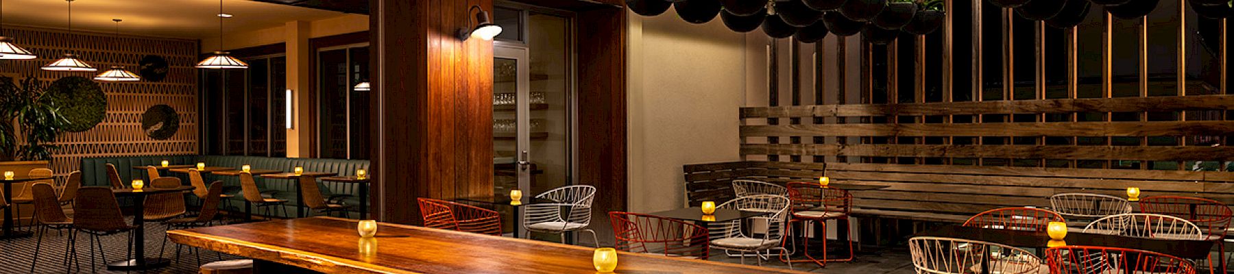 A cozy, dimly lit restaurant features wooden tables, stools, colorful chairs, hanging plants, and warm lighting throughout the space.