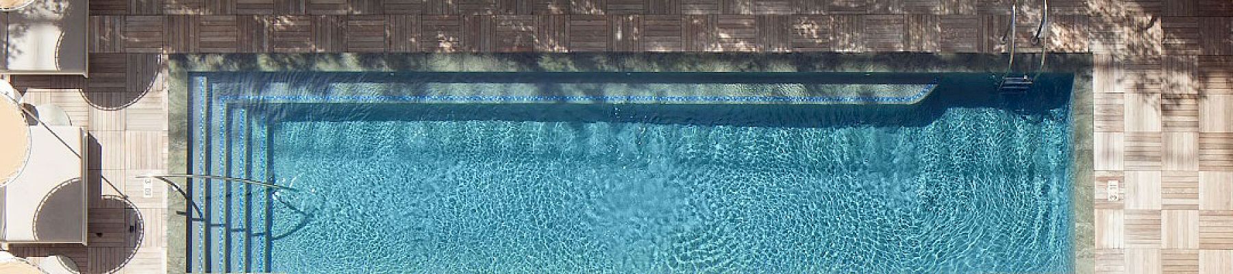 An aerial view of a pool with white sun loungers, plants surrounding, and seating areas on both sides, bordered by greenery.