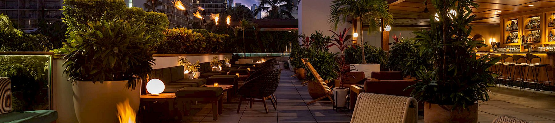 An outdoor lounge with a fire pit, cozy seating, and ambient lighting, set against an urban backdrop at dusk.